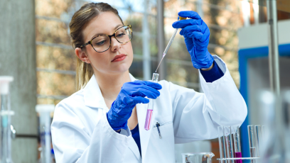 An image of a woman working in a lab wearing glasses, white lab coat, and gloves holding a beaker and dropper 