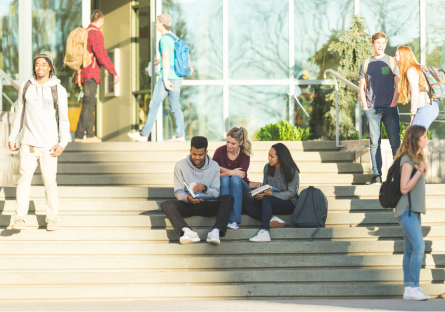 An image of college students sitting on steps in front of a campus building with other students walking nearby