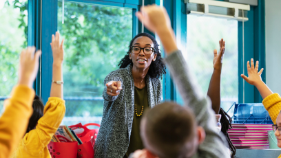 A stock image of a woman teacher pointing to students in a classroom with their hands raised.