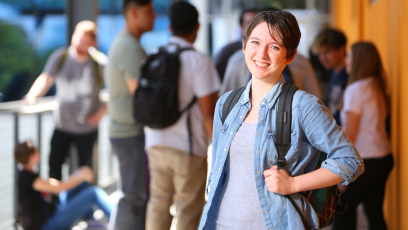 An image of a female college smiling with a backpack on one shoulder with other students in the background.