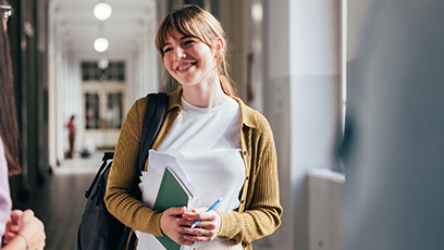 Image of female student holding text book while smiling in a hallway.