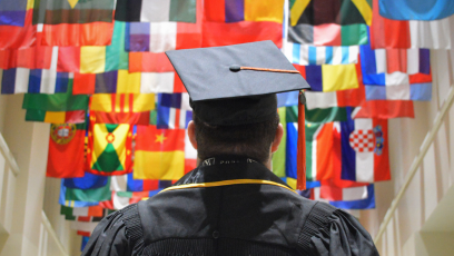 A college graduate  wearing a black cap and gown with back to camera looking at international flags