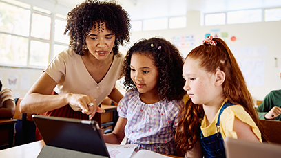 Image of a female teacher pointing to a tablet device while assisting two female school children in a classroom.