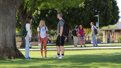 Three MCC students standing outside under a tree with three additional students in the background.