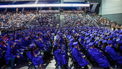 College graduates wearing royal blue caps and gowns seated in an auditorium.