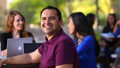 Image of a male student smiling sitting by computer with other students in the room