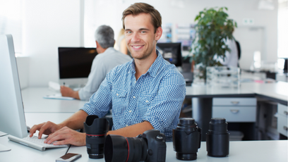 a man wearing a blue dress shirt sitting at a computer with camera equipment on the desk.