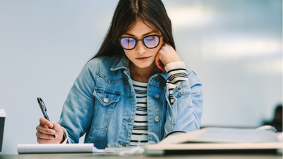 Image of a woman wearing glasses and a jean jacket holding a pen and reading a textbook and cup of coffee next to her