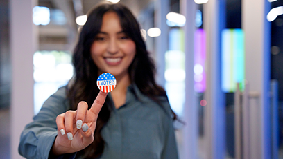 A woman smiling and holding her finger up that has a I Voted sticker on it.