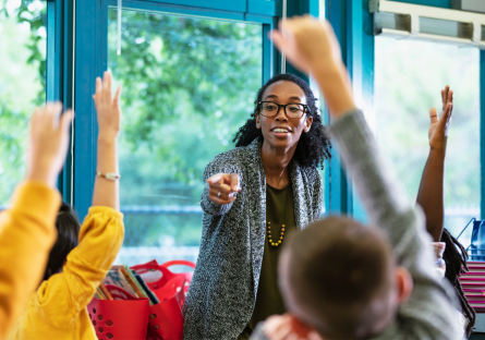 A stock image of a woman teacher pointing to students in a classroom with their hands raised.