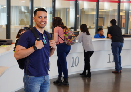 An image of a MCC student wearing a backpack standing in the college's enrollment center with students standing behind him at being helped by enrollment staff.