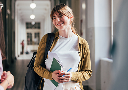 Image of female student holding text book while smiling in a hallway.