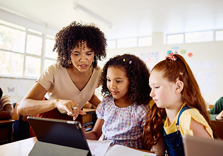Image of a female teacher pointing to a tablet device while assisting two female school children in a classroom.