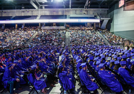 College graduates wearing royal blue caps and gowns seated in an auditorium.