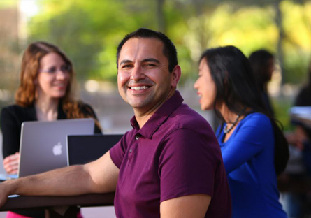 Image of a male student smiling sitting by computer with other students in the room