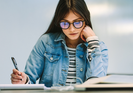 Image of a woman wearing glasses and a jean jacket holding a pen and reading a textbook and cup of coffee next to her