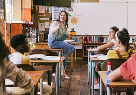 Teacher in classroom sitting on desk, speaking to group of students 
