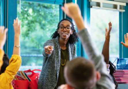 A stock image of a woman teacher pointing to students in a classroom with their hands raised.