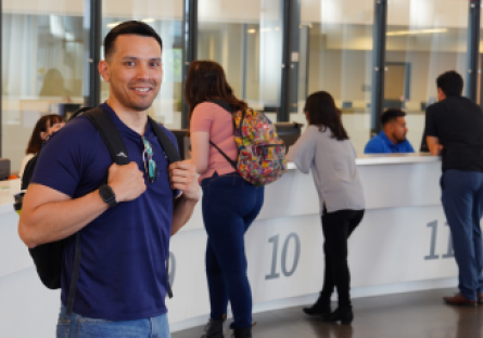 An image of a MCC student wearing a backpack standing in the college's enrollment center with students standing behind him at being helped by enrollment staff.