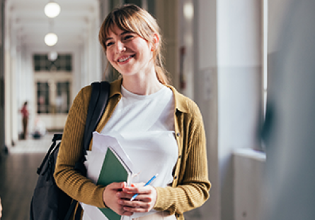 Image of female student holding text book while smiling in a hallway.