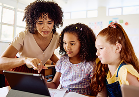 Image of a female teacher pointing to a tablet device while assisting two female school children in a classroom.