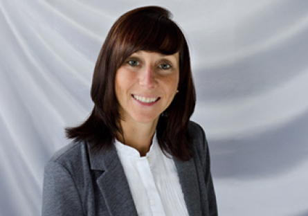 A gray background with a smiling Beth Roszatycki wearing a dark gray suit jacket and a white blouse.
