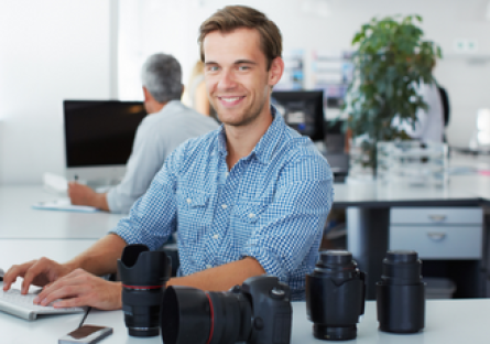 a man wearing a blue dress shirt sitting at a computer with camera equipment on the desk.