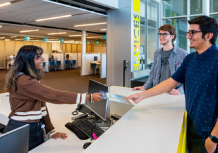 An image of a woman standing behind a desk assisting a make student while another male student looks on