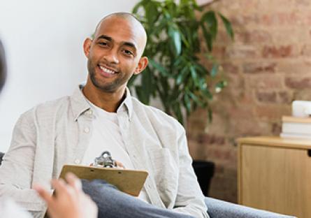 An image of a smiling male holding a clipboard listening to a woman