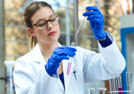 An image of a woman working in a lab wearing glasses, white lab coat, and gloves holding a beaker and dropper 