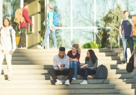 An image of college students sitting on steps in front of a campus building with other students walking nearby