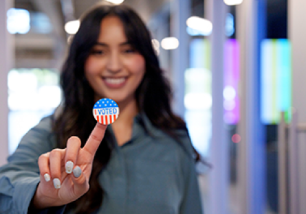 A woman smiling and holding her finger up that has a I Voted sticker on it.